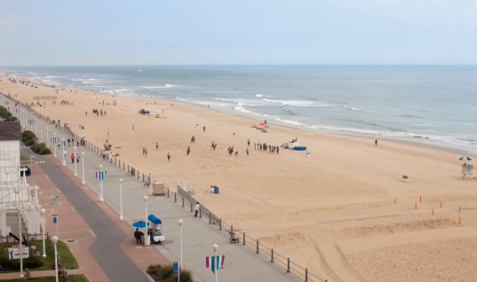 Shore scene from Virginia Beach.Shot from a high elevation, this shot is taken down the boardwalk and beach early in the morning. There are a few people there, but it is a calm and peaceful morning. The calm waves crash on the beach and it is a cloudy day and the boardwalk has a diminishing perspective.- A great beach shot taken in the outdoors.