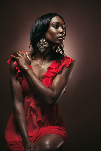 An attractive young adult African model poses for a portrait in a fashionable red evening dress and earrings.  Vertical studio shot on brown background