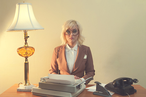 A retro styled photograph of a woman working in an office as a secretary or receptionist, complete with typewriter, rotary phone and kitsch desk lamp.  She looks bored.  Vertical with copy space.