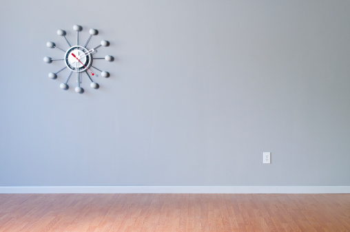Retro wall clock hanging in empty room with grey wall and wood flooring.