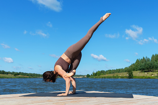 Woman doing Eka Pada Bakasana exercise, crane pose with leg extended up, balance asana, full length handstand, practicing yoga in sportswear on lakeside