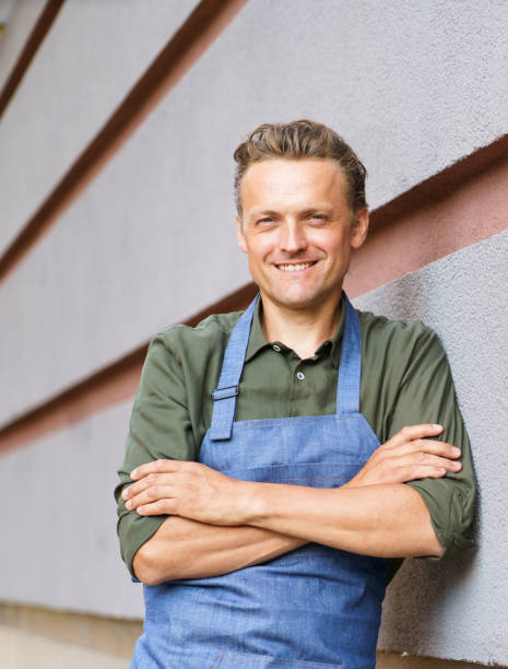 istock Skilled and dedicated waiter or service man in restaurant setting. Dressed in neat apron and uniform, he stands by the wall with crossed hands, radiating confidence and professionalism. 1576227554
