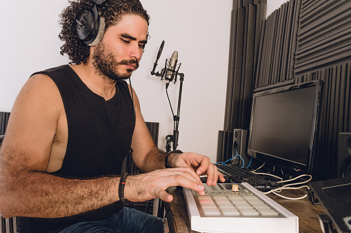 young latino man with curlers is sitting working in his home studio mixing a music production with a MIDI controller