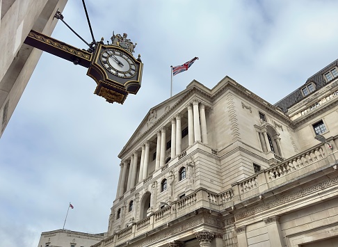 Close-up on a sign for the Bank of England, with pedestrians passing the institution's main entrance in the background.