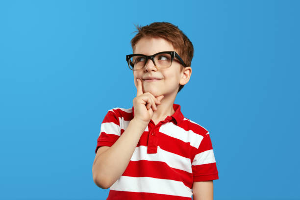 istock Thoughtful boy in nerdy glasses and red striped shirt touching cheek and looking up while thinking 1576308014