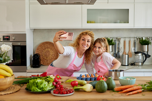 Beautiful woman and her cute daughter taking selfie in kitchen while preparing food