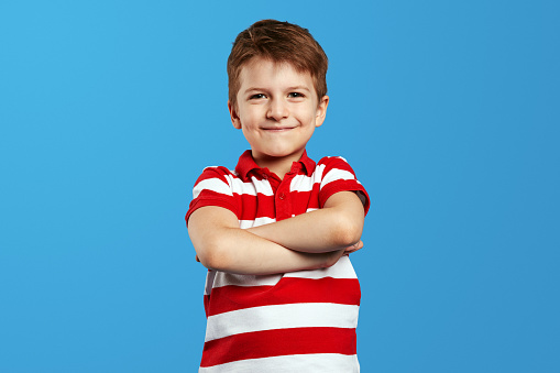 Little cute boy in red stripped shirt crossing arms and looking at camera while standing against blue background.