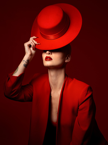 Hat, fashion and a model woman on a red background in studio for elegant or trendy style. Aesthetic, art and beauty with a confident young female person posing in an edgy, classy or unique suit