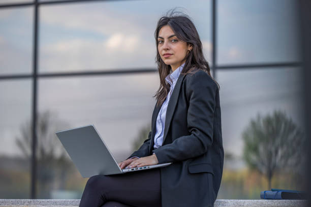 istock Serious business woman working hard and solving business problem on her laptop. She is sitting outdoors near modern office center. 1576343111