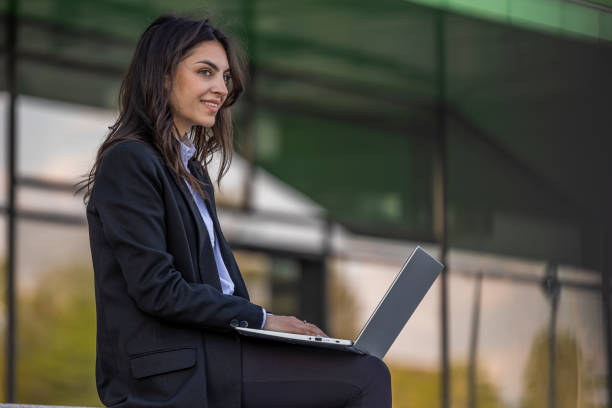 istock Smiling businesswoman working on laptop. Insurance agent checking email on computer and  sitting outdoors near modern office center. 1576344630