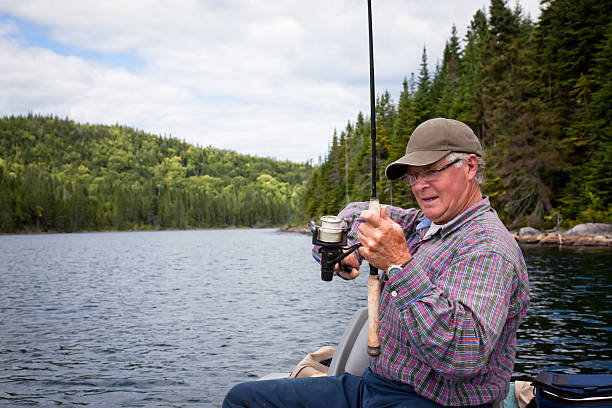 DSLR photo of a senior fisherman. The fisherman is sitting in a green motor boat and fishing. The lake is calm. Some mountains and trees are in the background of the picture. It is mostly a cloudy day with parts of blue sky. 