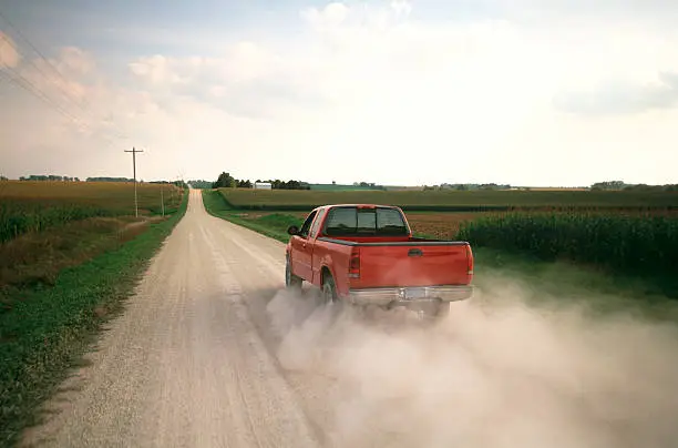 Red pick up truck traveling down dusty rural road.