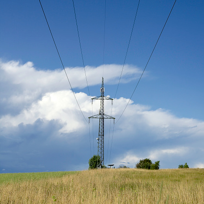 High voltage power lines and pylons in a flat green and partly plowed agricultural landscape, on a sunny day with clouds in the blue sky.