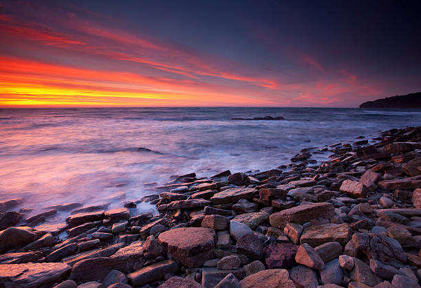 The sun rises over the sea near Bridport in Dorset, England. The Isle of Portland is just visible on the horizon. There is lots of motion blur in the foreground waves.