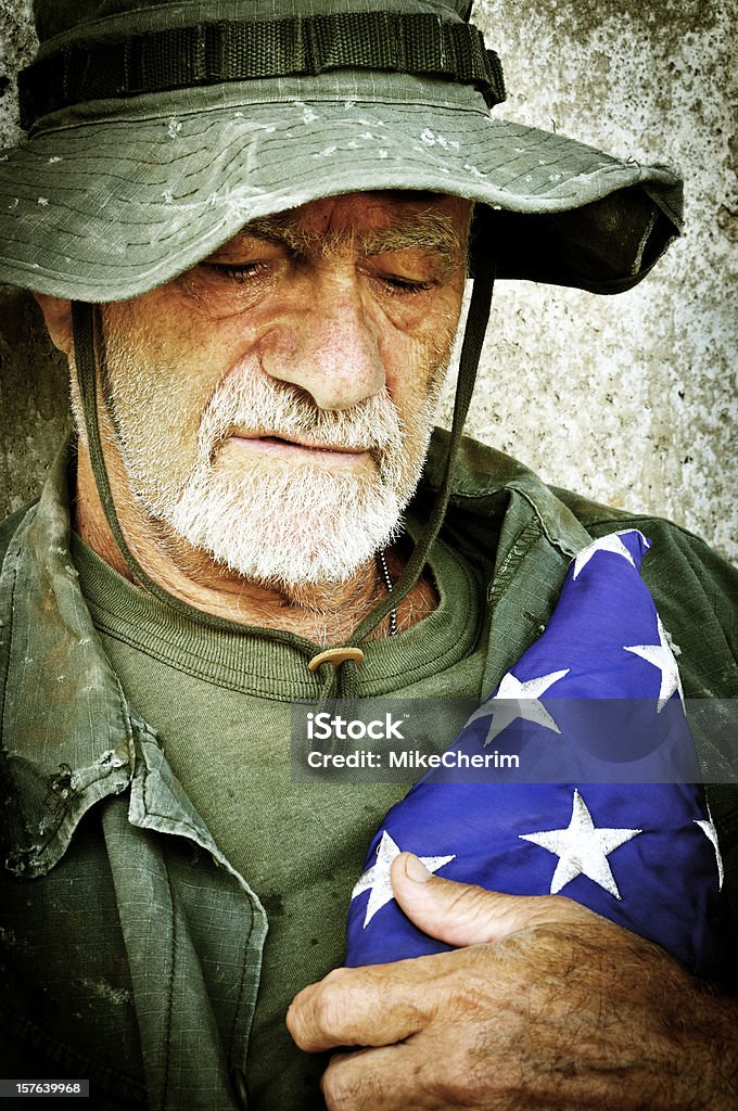 Vietnam Vet with Folded Flag Recalls the War (Xpro) A Vietnam war veteran (actual) appears to be down and out. In this shot, sad, he looks down, recalling the war, tightly hugging a folded flag, remembering his fallen brothers. His eyes are moist and has tears on his cheeks. Scene staged.  Veteran Stock Photo