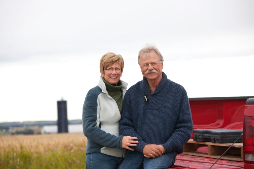 Happy farm couple with pickup truck and farm buildings in background.
