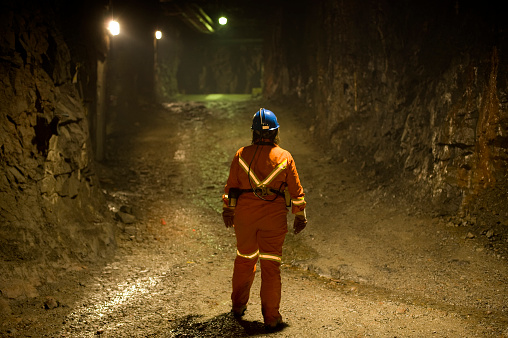 A woman mine worker in a mine shaft or tunnel underground.  She is looking away down the tunnel and wearing work overalls and safety gear.
