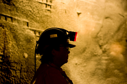 A woman working underground in a mine is silhouetted against a well lit tunnel wall.  She wears a hard hat helmet with a miners lamp on it.  Northwest Territories, Canada.