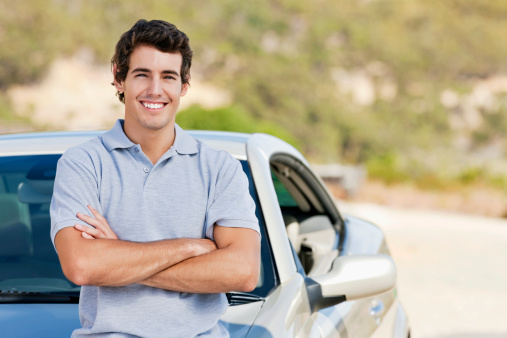 A young man smiles while standing in front of his car with his arms crossed.  Horizontal shot.