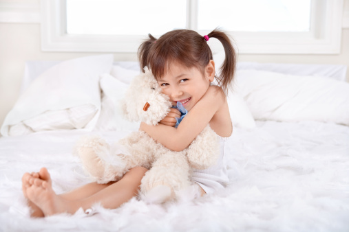 Adorable little Asian girl playing teddy bear while sitting on the bed at home.