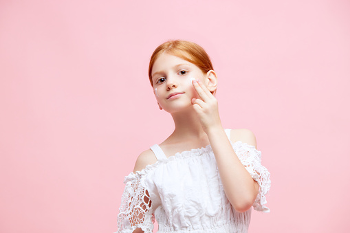 Portrait of little redhead girl, child applying face moisturizing cream against pink studio background. Concept of skincare, childhood, cosmetology and health, beauty, organic products, ad