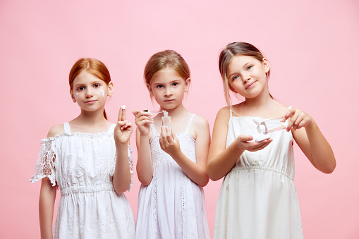 Pretty little girls, children using natural cosmetological face products for kids against pink studio background. Concept of skincare, childhood, cosmetology and health, beauty, organic products, ad