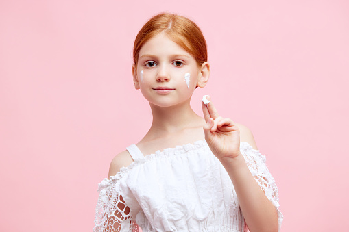 Portrait of little redhead girl, child applying face moisturizing cream against pink studio background. Concept of skincare, childhood, cosmetology and health, beauty, organic products, ad