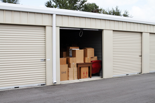 Warehouse building with self storage units. Self storage facility. Roll up doors on self storage facility. One door open with boxes, household goods and furniture in doorway. Several closed units also visible. 