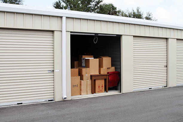 Self storage warehouse with single storage unit open to Warehouse building with self storage units. Self storage facility. Roll up doors on self storage facility. One door open with boxes, household goods and furniture in doorway. Several closed units also visible.  storage compartment stock pictures, royalty-free photos & images