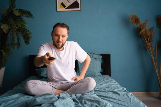 istock Sleepless man in pajama watching tv while sitting on bed with remote controller 1576425559