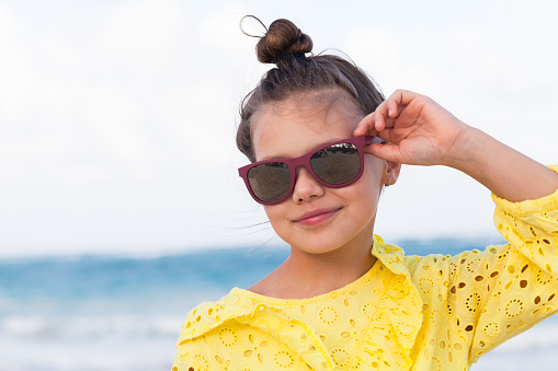 Happy little girl in yellow dress and sunglasses stands on a beach on a sunny summer day. Closeup portrait