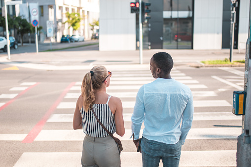 Behind the back shot of a diverse businesswoman and businessman standing at a traffic light before crossing the street in a business district. Both wearing smart casual clothes.