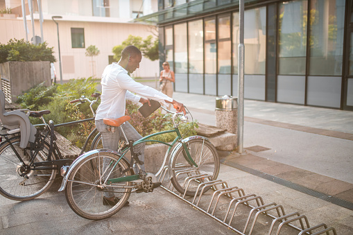 Black businessman arriving to work by bicycle. Parking it in front of a corporate building. He is wearing smart casual clothes.