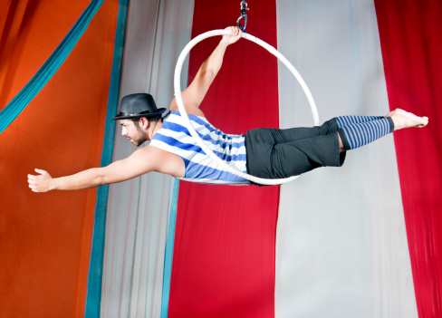 adult man doing tricks in a circus ring dressed with black pants and hat and blue and white stripes on his tank top with a colorful background