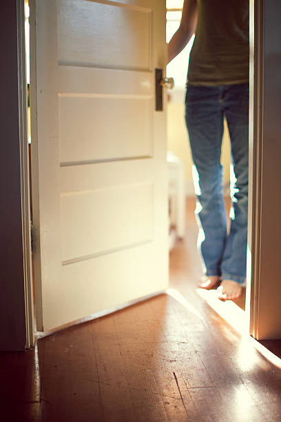 A young adult stands behind a partially closed door, sunlight streaming through from behind.  Focus on the floor in foreground.  Vertical with copy space.