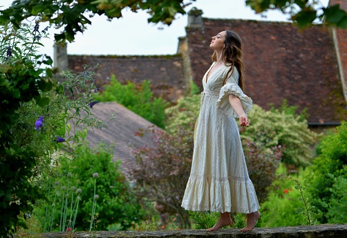 Young elegant brunette woman in white vintage dress raising her arms to the sky, facing the green nature and the typical roofs of the Dordogne, France, Europe