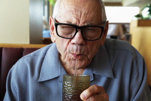 A senior man sitting in a diner-style restaurant squints as he drinks from a water glass through a straw. Selective focus on his face.