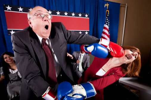 Two mature politicians (one male, one female) engage in a boxing match on stage instead of resolving their differences amicably.