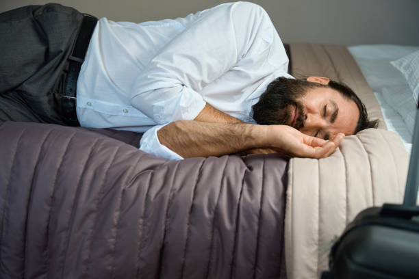 Tired male with a headache lay down to rest on the bed in a hotel room, next to travel suitcase