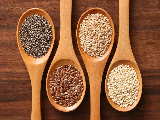 Four light-colored wooden spoons filled with different types of seeds positioned on a dark wood table. From left to right, the spoons contain chia, flax, sesame and quinoa seeds.  They are arranged with the handle facing in the opposite direction with every other spoon.