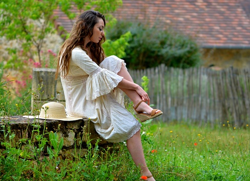 Young elegant brunette woman in vintage white dress and straw hat sitting on a low wall in a bucolic setting changing shoes, France, Europe