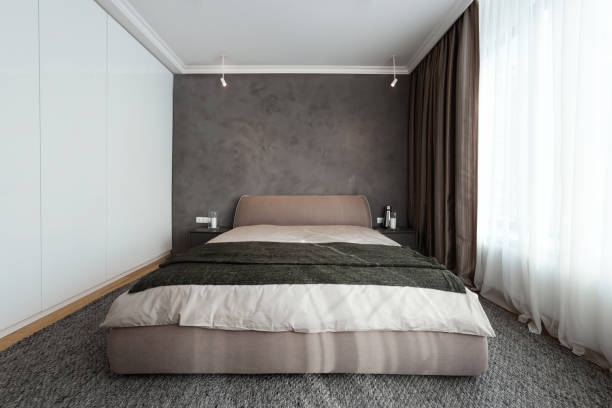 Light bedroom with double bed and window in hotel room. Well furnished space with comfortable sleeping zone, bedside table and built-in wardrobe on grey carpet