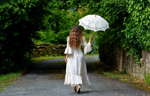 Young elegant brunette woman in vintage white dress and white parasol seen from the back, walking on a path, Dordogne, France, Europe