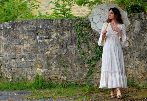 Young elegant brunette woman in white vintage dress and white parasol standing against a stone wall in a bucolic and green setting, Dordogne, France, Europe
