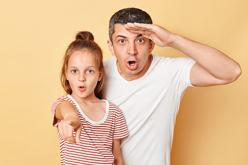 Shocked father with hand near forehead and daughter pointing at camera family wearing casual t-shirts standing isolated over beige background.