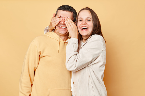 Happy optimistic woman and man wearing casual style clothing standing isolated over beige background couple having fun wife covering husband eyes.