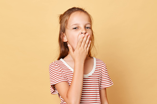 Tired little brown haired girl wearing striped t-shirt standing isolated over beige background covering mouth with palm yawning felling sleepy.
