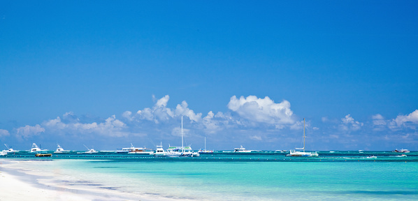A panoramic photo of a tropical beach with white sand and turquoise water. The sky is blue with a few white clouds. There are several boats and yachts in the water. The water is calm and clear, and you can see the ocean floor through it. The beach is empty and there are no people visible in the photo. This image can be used for travel, vacation, relaxation, or nature themes.
