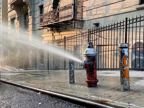 Open red and silver fire hydrant fitted with a city-approved spray cap that conserves water sprinkling water onto the street on a hot summer evening in Harlem, New York City