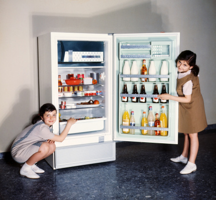 Two smiling girls proudly posing by an open refrigerator. All products in the fridge altered to avoid copyright issues. Sixties film scan, some grain visible.